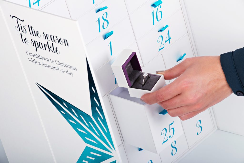 A hand reaching into a white and blue advent calendar to take out a diamond ring in a box