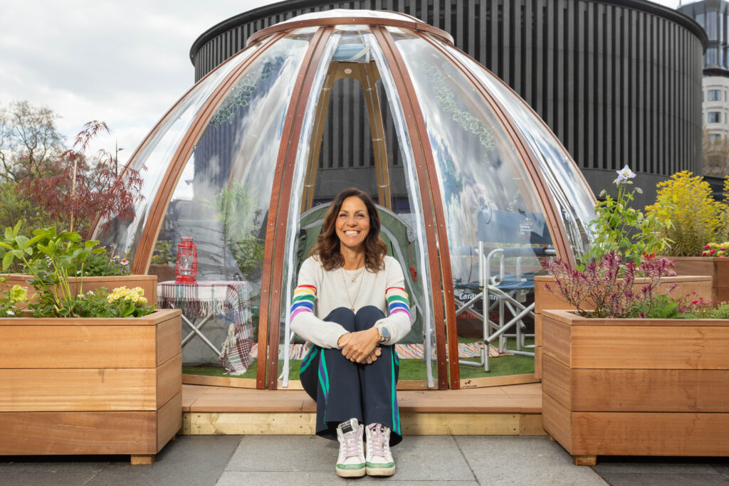Julia Bradbury sitting in the doorway of a see-through camping pod filled with plants