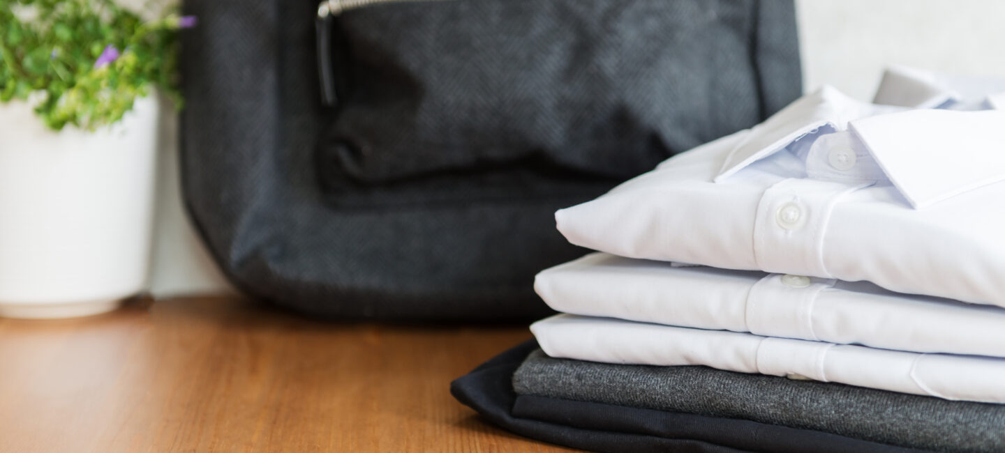 A pile of white school shirts and grey and black trousers alongside a black backpack on a wooden table