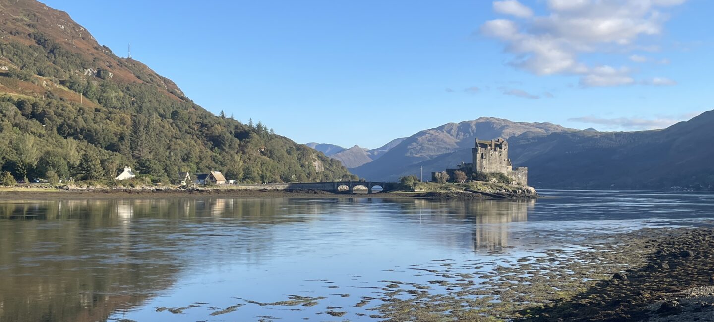 Landscape image of a castle-type building on a lake set against surrounding hills under a blue sky