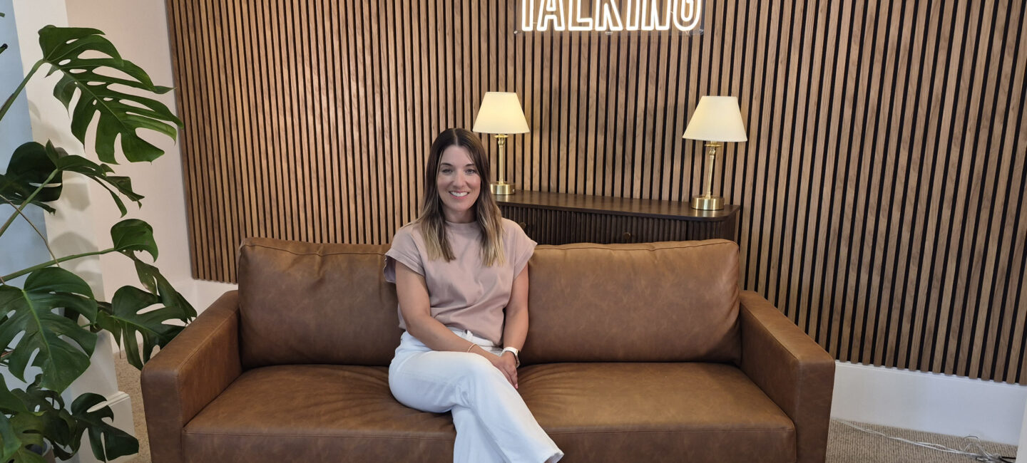 Bex Williams sitting on a brown sofa in front of a wood panelled wall, smiling